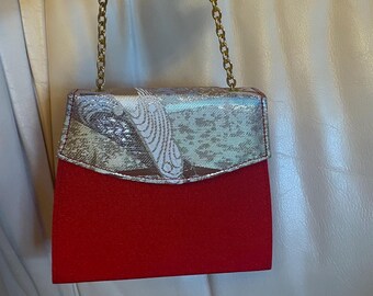 Könnte beinhalten: Kleine, rechteckige rote Handtasche mit goldenem Kettenriemen. Die obere Klappe ist silber- und goldfarben mit einem gemusterten Design. Die Tasche wird von einer Hand gehalten.
