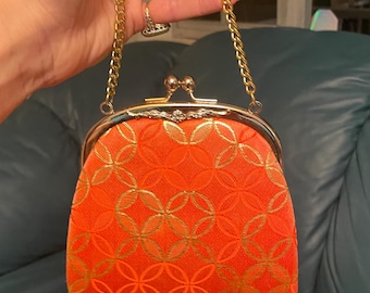Könnte beinhalten: Orangefarbene Clutch mit goldener Kette. Die Handtasche hat einen goldenen Verschluss und ein sich wiederholendes goldenes Kreismuster auf der Vorderseite. Die Tasche wird von einer goldenen Kette gehalten.