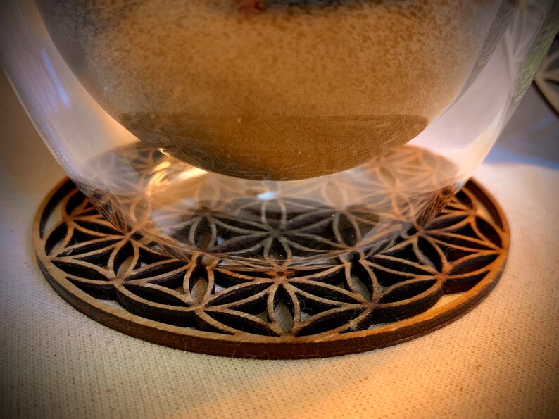 May include: A wooden coaster with a geometric pattern, resembling a flower of life design, is shown with a clear glass cup on top.
