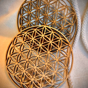 May include: Two wooden coasters with a geometric flower of life design. The coasters are round and have a light brown color.