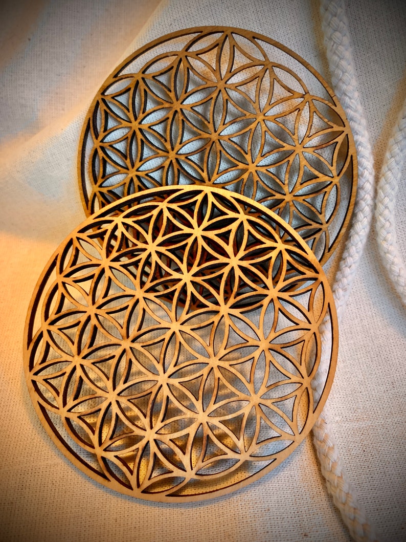 May include: Two wooden coasters with a geometric flower of life design. The coasters are round and have a light brown color.