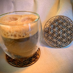 May include: A glass of coffee with foam on top, sitting on a wooden coaster with a geometric pattern. A second coaster with the same pattern is to the right of the glass.