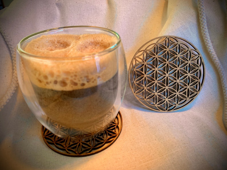 May include: A glass of coffee with foam on top, sitting on a wooden coaster with a geometric pattern. A second coaster with the same pattern is to the right of the glass.