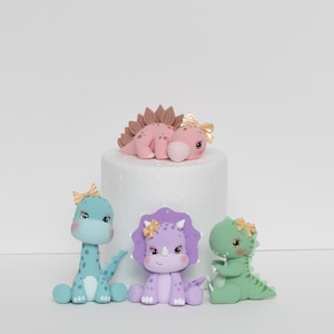 May include: Four pastel-coloured dinosaur cake toppers. A pink dinosaur with brown spikes lies on a white cake stand. A light blue dinosaur with a pink bow sits on the left. A purple dinosaur with a gold bow sits in the centre. A green dinosaur with a gold bow sits on the right.