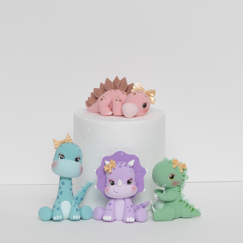 May include: Four pastel-coloured dinosaur cake toppers. A pink dinosaur with brown spikes lies on a white cake stand. A light blue dinosaur with a pink bow sits on the left. A purple dinosaur with a gold bow sits in the centre. A green dinosaur with a gold bow sits on the right.