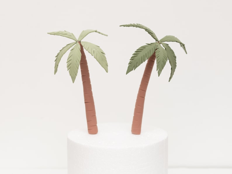 May include: Two green palm tree cake toppers with brown trunks. The palm trees are made of fondant and are perfect for a tropical themed cake.