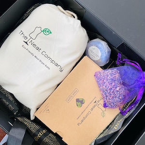 May include: A black gift box with a white bag, a brown box, a purple bag filled with lavender, and a lavender bath bomb. The white bag has the text "The Neat Company Eco-Friendly Wool Dryer Balls". The brown box has the text "The Neat Company Eco-Friendly Laundry Detergent Sheets Fresh Scent". The bath bomb has the text "The Neat Company Eco-Friendly Laundry Detergent Sheets Lavender".