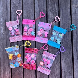 Può includere: Sette piccole bustine di plastica trasparente piene di ciondoli in miniatura colorati, ciascuna con un portachiavi a forma di cuore. I ciondoli includono varie forme come ciambelle, stelle e animali. Le bustine hanno bordi decorativi.