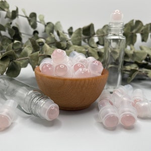 May include: A bowl filled with pink rollerball bottles, with a few more rollerball bottles scattered on a white surface. The rollerball bottles are made of clear glass with a pink stone rollerball.