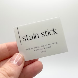 Op de afbeelding: Witte vlekken-stick met zwarte tekst "stain stick" en gebruiksaanwijzing: "roll on stain, let sit for 10-20 minutes & wash 30 ml".