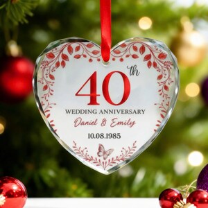 May include: Heart-shaped glass ornament with a red ribbon for hanging. The ornament features the number "40" in red, the words "Wedding Anniversary", names "Daniel & Emily", and the date "10.08.1985". Decorative red floral elements and a butterfly are also present.
