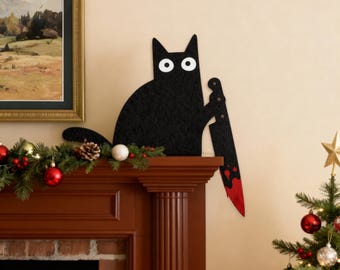 May include: Black cat-shaped wall decoration holding a bloody knife. The cat has white eyes and is made of a textured material. The decoration is placed above a fireplace decorated with Christmas ornaments and a Christmas tree.