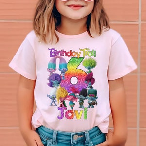 May include: A light pink t-shirt featuring a colorful graphic of Trolls characters, the number 6, and the text "Birthday Troll" and "Jovi". The shirt is worn by a child wearing sunglasses and a hat.