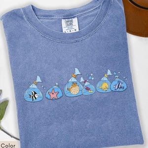May include: Blue jean colored t-shirt with a crew neck. The shirt features a graphic of several cartoon fish in plastic bags. The fish include a black and white striped fish, a pink starfish, and a yellow fish. The shirt has the text "COMFORT COLORS" on the tag.