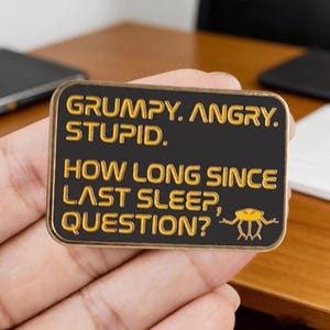 May include: A rectangular black pin with gold lettering that reads "GRUMPY. ANGRY. STUPID." and "HOW LONG SINCE LAST SLEEP, QUESTION?" A small yellow insect graphic is at the bottom. The pin is held in a hand.