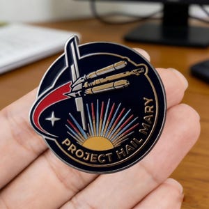 May include: A circular, navy blue metal pin with a gold border. The pin features a spaceship design with the text "PROJECT HAIL MARY". The design includes a star, a rising sun, and a red and white trail.