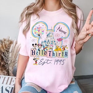Könnte beinhalten: Rosa T-Shirt mit einem Aufdruck von Mickey Mouse und seinen Freunden in Disney World. Der Aufdruck zeigt ein Schloss, Ballons und den Text "Hello Thirty Est. 1995".