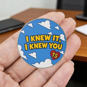 May include: A circular enamel pin featuring a bright blue sky background with white clouds. The text "I KNEW IT, I KNEW YOU" is printed in bold yellow letters. A small red heart icon with the letters "TS" in yellow is positioned at the bottom right.
