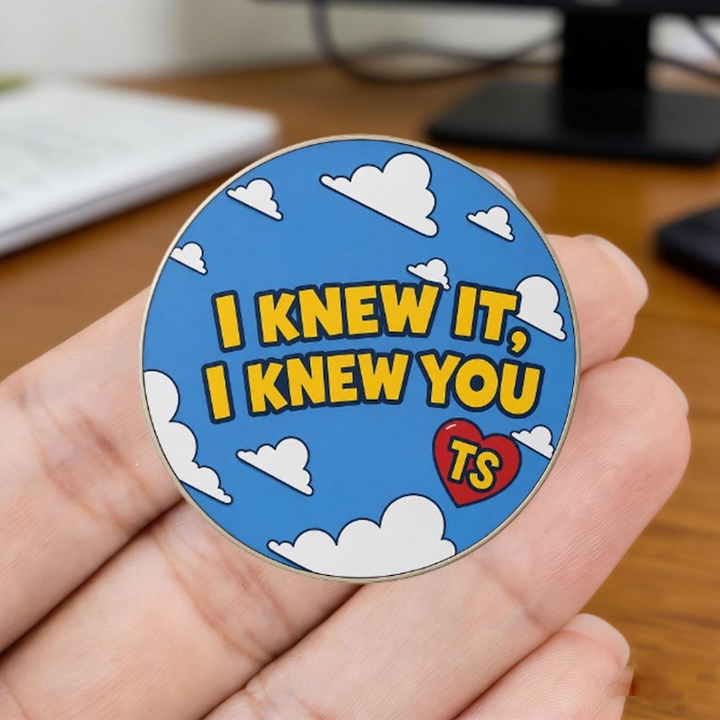 May include: A round enamel pin featuring a blue sky background with white clouds. The text "I KNEW IT, I KNEW YOU" is printed in bold yellow letters. A small red heart icon contains the yellow letters "TS" in the lower right corner of the pin.