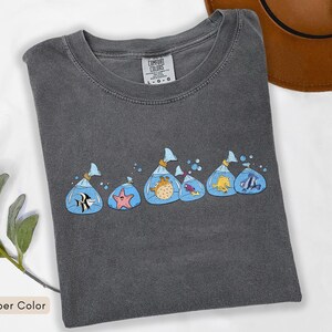 May include: A dark grey Comfort Colors t-shirt with a design of cartoon fish in plastic bags. The fish are in various colours and species, with blue water and bubbles. The shirt is a pepper colour.