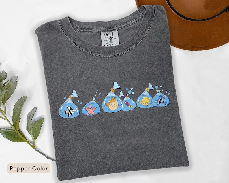 May include: A dark grey Comfort Colors t-shirt with a design of cartoon fish in plastic bags. The fish are in various colours and species, with blue water and bubbles. The shirt is a pepper colour.