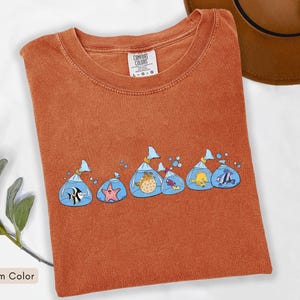 May include: A yam-coloured t-shirt with a design of cartoon fish in plastic bags. The fish are in various colours and species, with blue water and bubbles. The shirt has a crew neck and short sleeves. The tag reads "Comfort Colors".