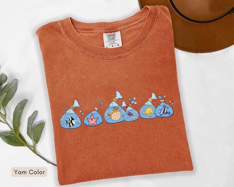 May include: A yam-coloured t-shirt with a design of cartoon fish in plastic bags. The fish are in various colours and species, with blue water and bubbles. The shirt has a crew neck and short sleeves. The tag reads "Comfort Colors".