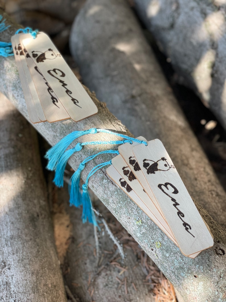 May include: A set of four wooden bookmarks featuring a panda bear design and the name "Emea" inscribed on each. The bookmarks have blue tassels.