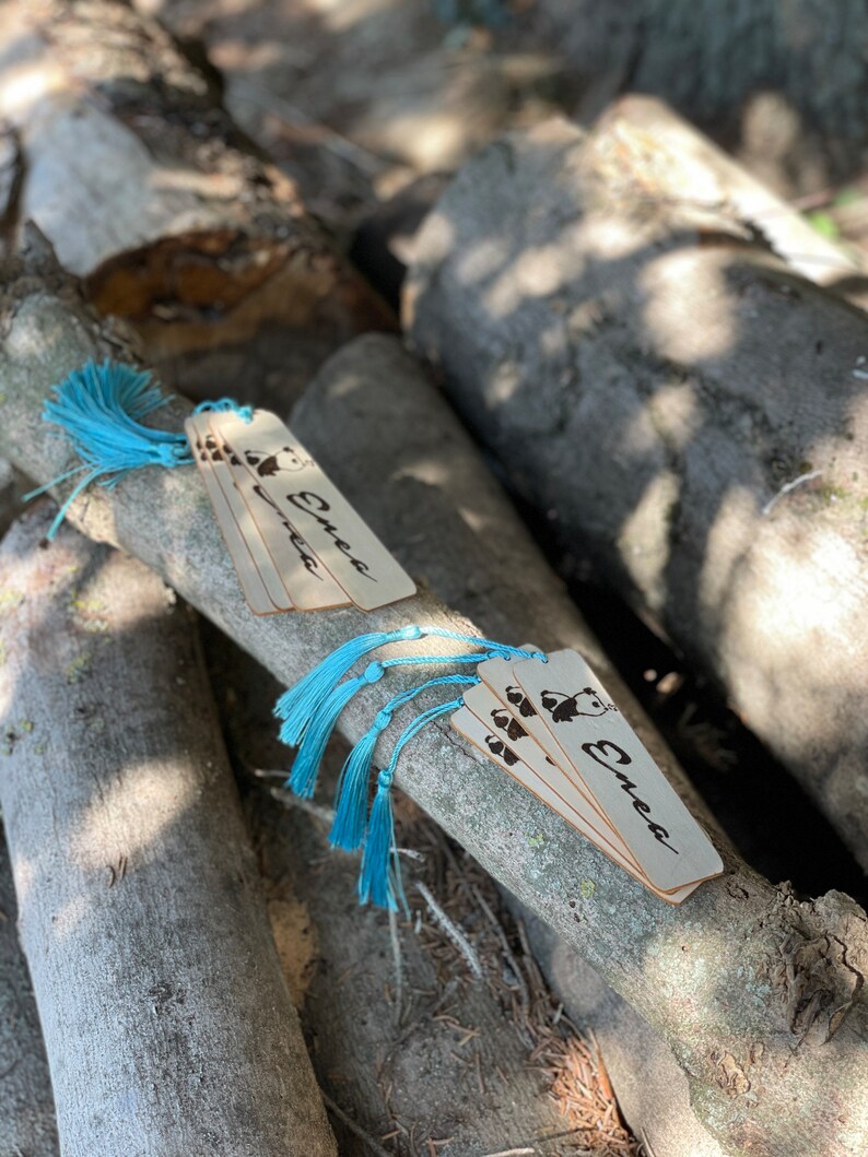 May include: A set of five wooden bookmarks featuring a panda bear design and the name "Enea" inscribed on them. Each bookmark has a blue tassel attached.
