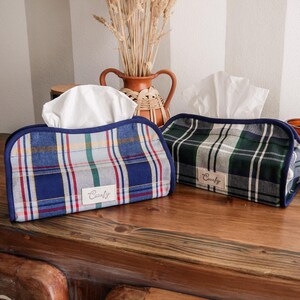 Puede incluir: Dos fundas para cajas de pañuelos a cuadros, una azul y otra verde, sobre una superficie de madera. La funda azul tiene detalles rojos, blancos y verdes, mientras que la verde tiene detalles blancos y grises. Ambas tienen una etiqueta "Comfy".