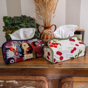 May include: Two decorative tissue box covers. One has a colourful abstract design with a blue border, the other has a white background with red cherries and a green border. Both have the word "Comfy" on them.
