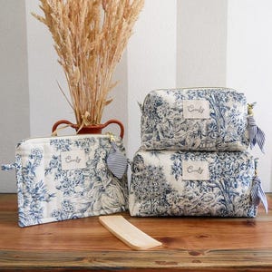 Può includere: Tre trousse per cosmetici di diverse dimensioni, con un design pastorale blu e bianco e la parola "Comfy". Le borse hanno cerniere dorate e nappe a righe. Sono disposte su una superficie di legno, con un vaso di piante essiccate.