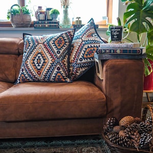 Peut inclure: Un canapé en cuir marron avec deux coussins décoratifs à motifs. Les coussins présentent un motif géométrique dans des tons de bleu, orange, rouge et blanc. Le canapé est devant une fenêtre avec vue sur une plante verte et un vase de fleurs séchées. Il y a une tasse à café sur une table d'appoint avec le texte "Premium Coffee" dessus.