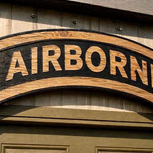 May include: Wooden sign with the word "AIRBORNE" in large, bold, light-colored letters against a black background. The sign has a curved shape and is framed by a lighter wood border. The sign is mounted on a light-colored wall.