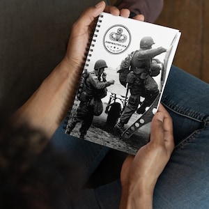 May include: Spiral-bound notebook featuring a black and white image of soldiers. The cover includes a circular emblem with the text "JUMPMASTER USA" and "UP TO NO GOOD". The notebook is held open, displaying the image of soldiers.