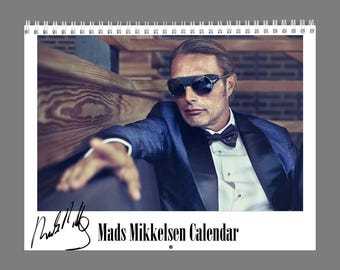 May include: A spiral-bound wall calendar featuring a photograph of Mads Mikkelsen wearing a blue tuxedo jacket, white dress shirt, black bow tie, and dark sunglasses. The bottom of the page displays the text Mads Mikkelsen Calendar and a signature.