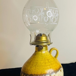 Puede incluir: Lámpara de aceite vintage con un globo de cristal transparente grabado con un diseño decorativo. La base es una jarra de cerámica redondeada con un esmalte amarillo y marrón, y un asa pequeña. El quemador es de latón.