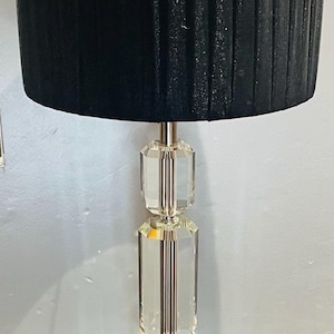 May include: A contemporary table lamp with a black, pleated lampshade. The lamp has a clear, faceted crystal base and a square, silver-toned metal base. Designed for indoor use, it includes a power cord.