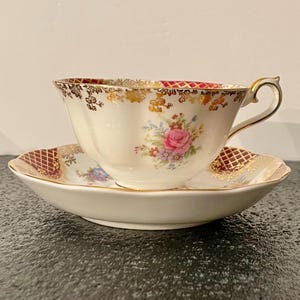 May include: A white teacup and saucer with a floral pattern and gold trim. The saucer has a red and gold chequered pattern.