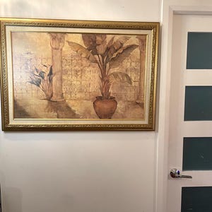 May include: A framed artwork featuring a sepia-toned illustration of a potted plant with large leaves, set against a backdrop of columns and a decorative wall. The artwork is enclosed in a gold-colored ornate frame, adding a touch of elegance.