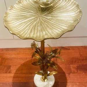 May include: A gold-coloured decorative side table with a lotus flower design. The table features a large, textured, leaf-shaped top and a smaller flower at the centre. The base is white marble with gold accents and botanical details.