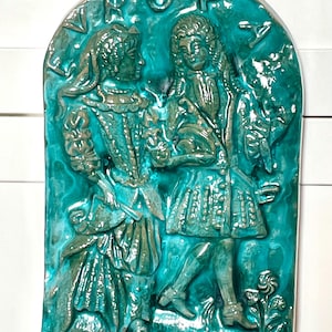 May include: A teal-colored ceramic wall plaque featuring a bas-relief of two figures in period clothing. The figures are facing each other, and the plaque has an arched top. The word "ROP" is at the top of the plaque. The figures are in a garden setting.