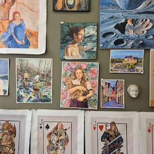 May include: A collection of various artworks, including paintings and prints, displayed on a wall. The pieces feature diverse subjects, such as portraits, landscapes, and fantasy scenes. Some artworks are framed, while others are unframed prints, including playing card designs.