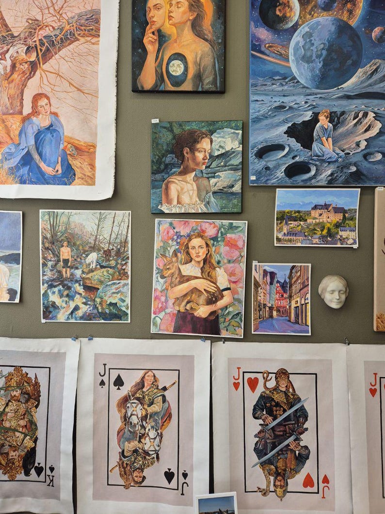 May include: A collection of various artworks, including paintings and prints, displayed on a wall. The pieces feature diverse subjects, such as portraits, landscapes, and fantasy scenes. Some artworks are framed, while others are unframed prints, including playing card designs.