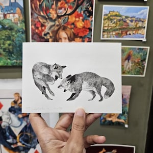May include: A monochrome ink drawing depicting two foxes in a dynamic interaction. The artwork is presented on a white rectangular card. The card is held in a hand, with other framed art pieces visible in the background. The foxes are the central subject.