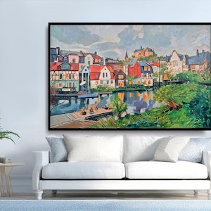 May include: A framed watercolor painting depicts a European townscape with colorful buildings along a river. The artwork features red-roofed houses, reflections in the water, and lush greenery, creating a serene landscape.