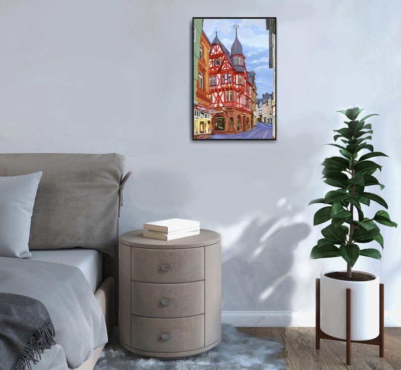 Pu&ograve; includere: Un dipinto incorniciato di una scena di strada europea con un edificio rosso e bianco. L'opera d'arte &egrave; esposta su una parete bianca sopra un comodino rotondo a tre cassetti. A destra c'&egrave; una pianta alta in vaso.