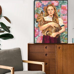 May include: A framed art print featuring a woman holding a fawn, set against a floral background of pink and purple blooms. The artwork is displayed on a wooden cabinet, with a potted plant and decorative items nearby. The overall aesthetic is warm and inviting.