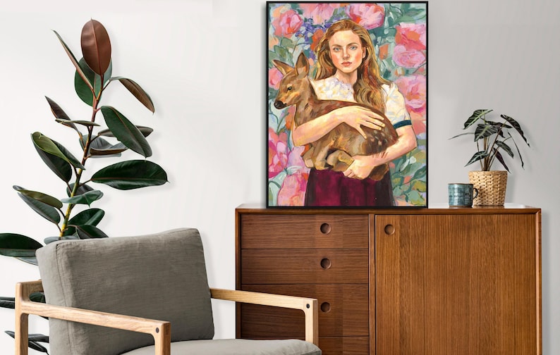 May include: A framed art print featuring a woman holding a fawn, set against a floral background of pink and purple blooms. The artwork is displayed on a wooden cabinet, with a potted plant and decorative items nearby. The overall aesthetic is warm and inviting.