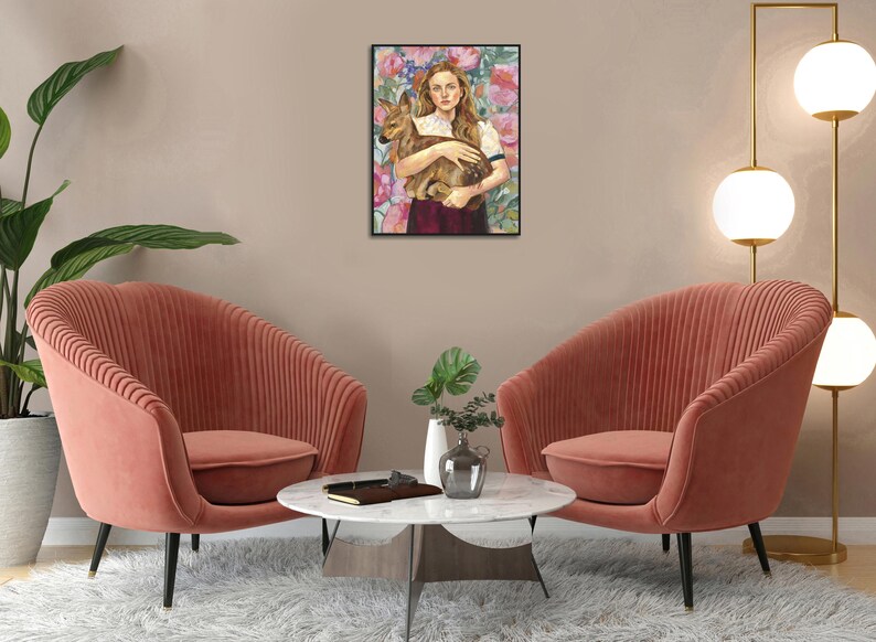 May include: A framed art print depicts a woman holding a fawn against a floral background. The artwork features a color palette of pink, brown, and white. Two coral-colored armchairs are in the foreground, with a white marble table and a gold lamp.